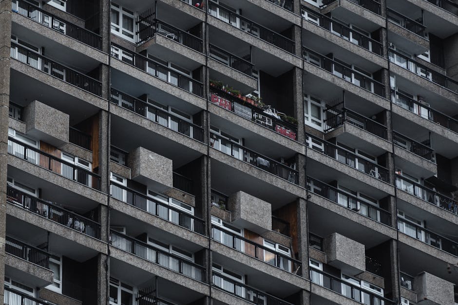 Close-up view of a residential apartment building façade featuring multiple concrete balconies with black metal railings. Some balconies have potted plants, laundry racks, and outdoor furniture, while