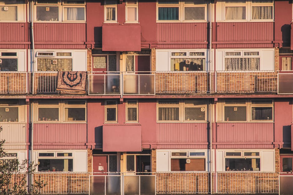 Close-up view of a residential apartment building façade featuring multiple concrete balconies with black metal railings. Some balconies have potted plants, laundry racks, and outdoor furniture, while